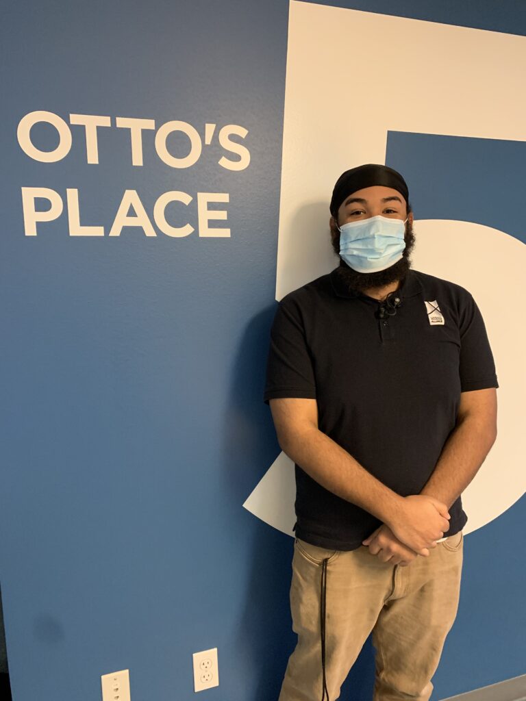 Ezekiel a housing navigator poses in front of the Otto's Place sign